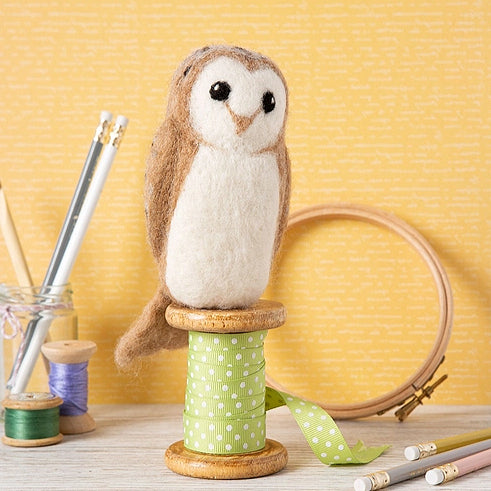 A felt owl figurine with brown and cream feathers sits on a green-and-white polka dot spool, styled with sewing and craft props against a yellow patterned backdrop.