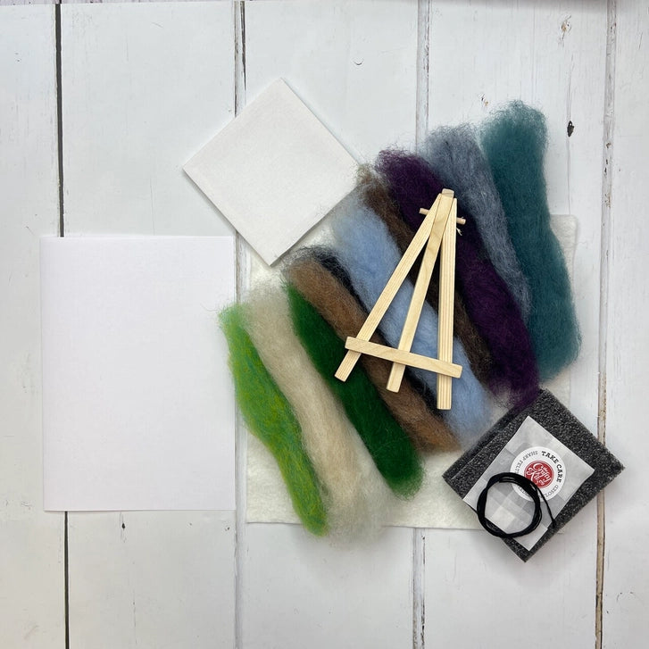 A flat-lay of a needlework kit on a white wood plank background, featuring wool fibres in green, beige, brown, light blue, purple, and teal, a small wooden easel, two white squares of fabric, a black felting pad.