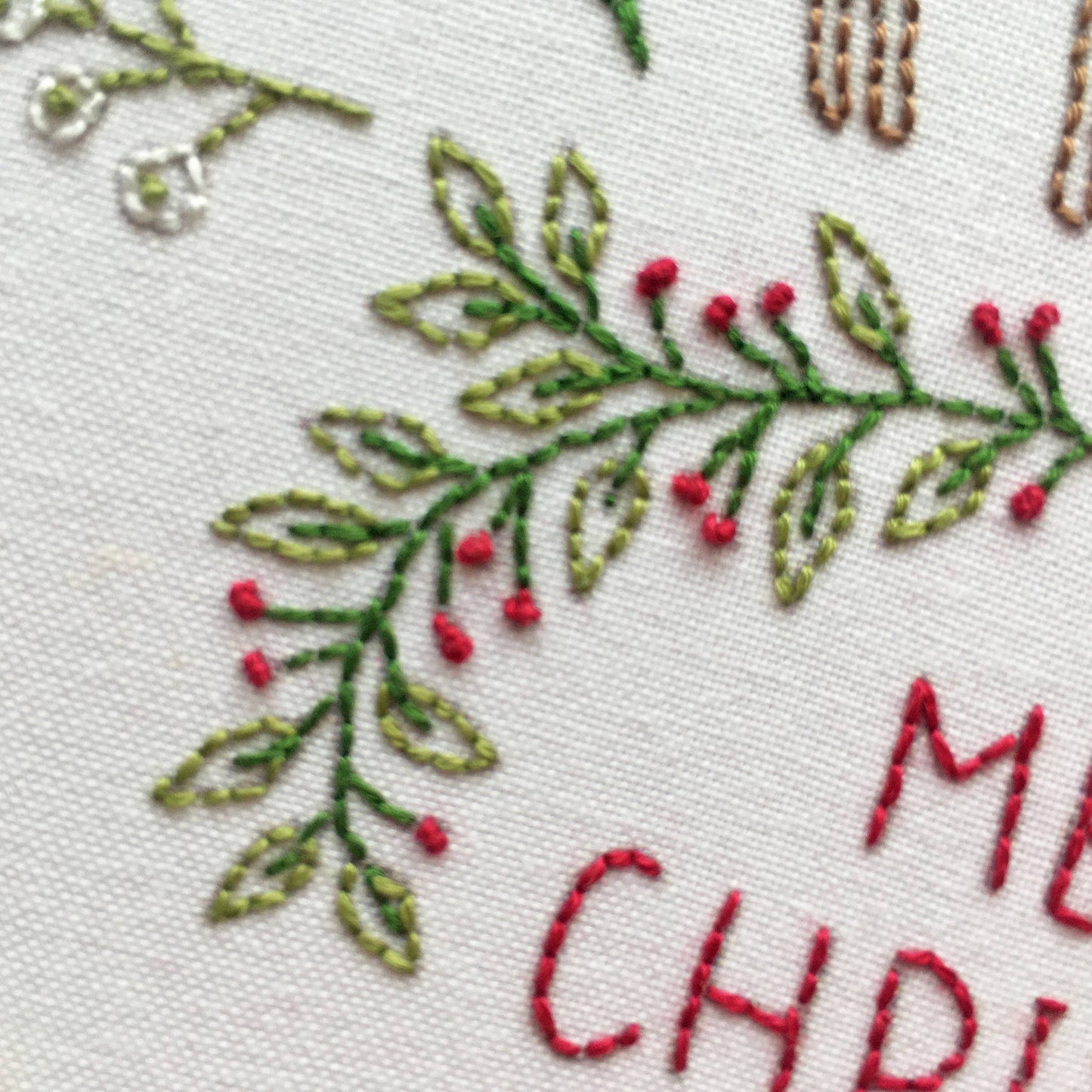 Close-up of embroidered holiday greenery with red berries on a white fabric background, with partial red embroidered text visible at the lower right.