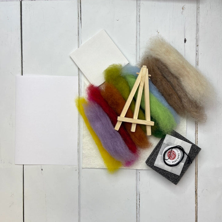 A flat-lay view of a wool felting kit on a white wood plank background, containing wool roving in various colours, a small wooden easel, felt cards and a felting block.
