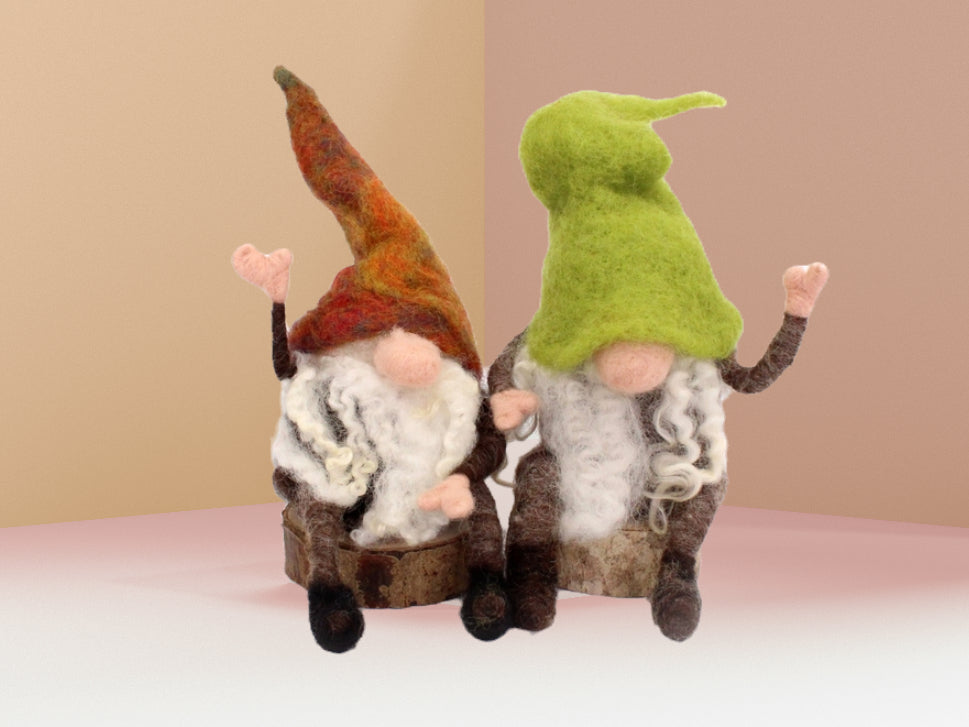 Two felted wool gnome figurines sitting side by side on a light pink surface against a beige background. The left gnome has a tall rust-orange hat, white beard, and dark brown body.