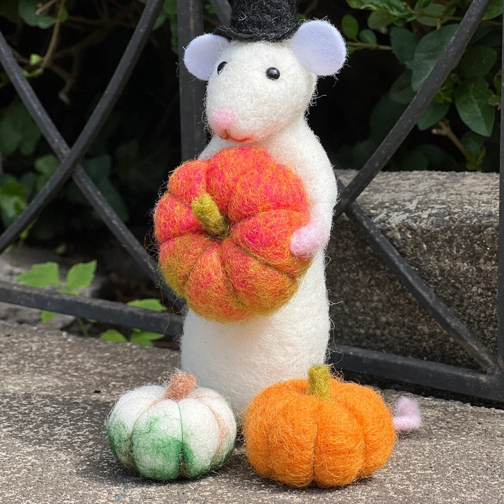 Felted mouse figurine with a hat holding a felt pumpkin, standing on a stone surface with more pumpkins around.