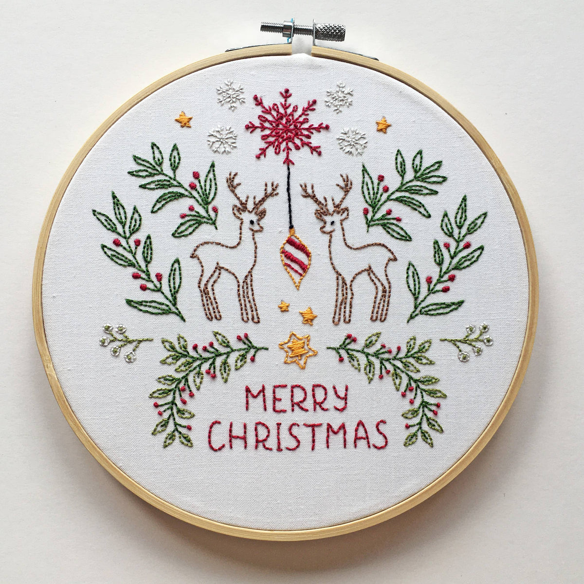 A round embroidery hoop holding a white fabric with a Christmas-themed stitched design. The design features two brown deer, a red ornament, green leafy branches with red berries, small gold stars, white snowflakes, and the words 'MERRY CHRISTMAS' in red.
