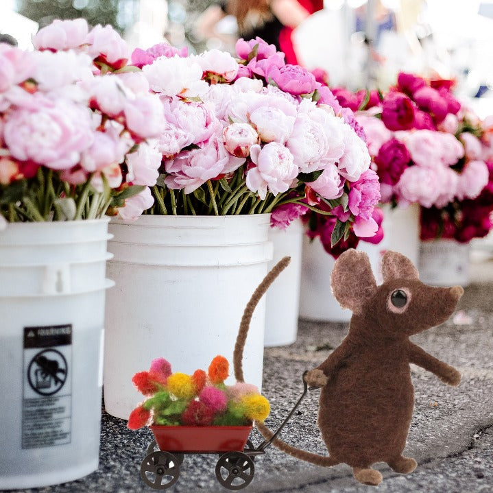 A small brown needle felted mouse toy pulls a red cart filled with flowers in front of white buckets of pink and magenta peonies.