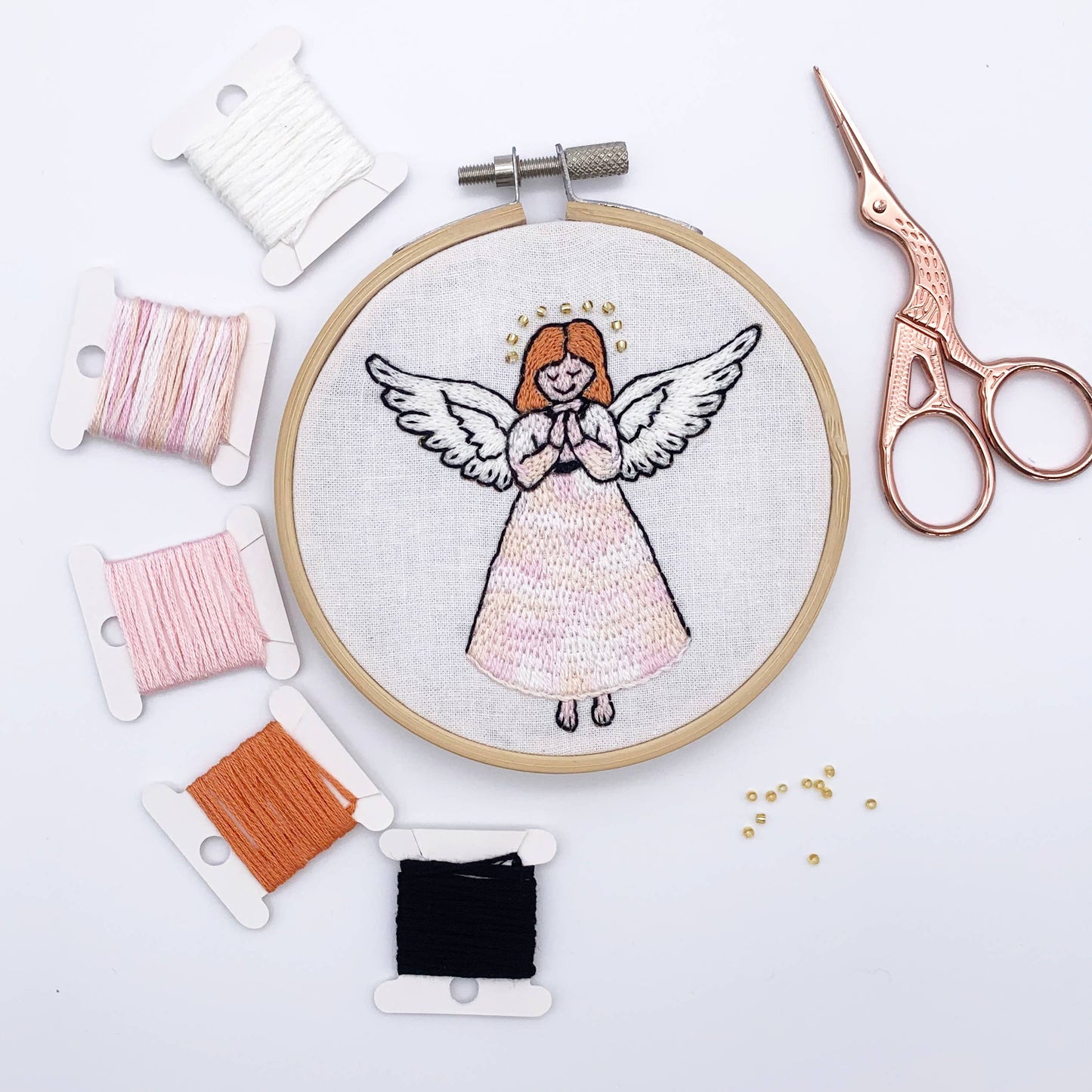 A circular embroidery hoop displaying a finished angel embroidery design on white fabric, surrounded by spools of thread in white, pink, orange, and black, with rose gold embroidery scissors on a white background.