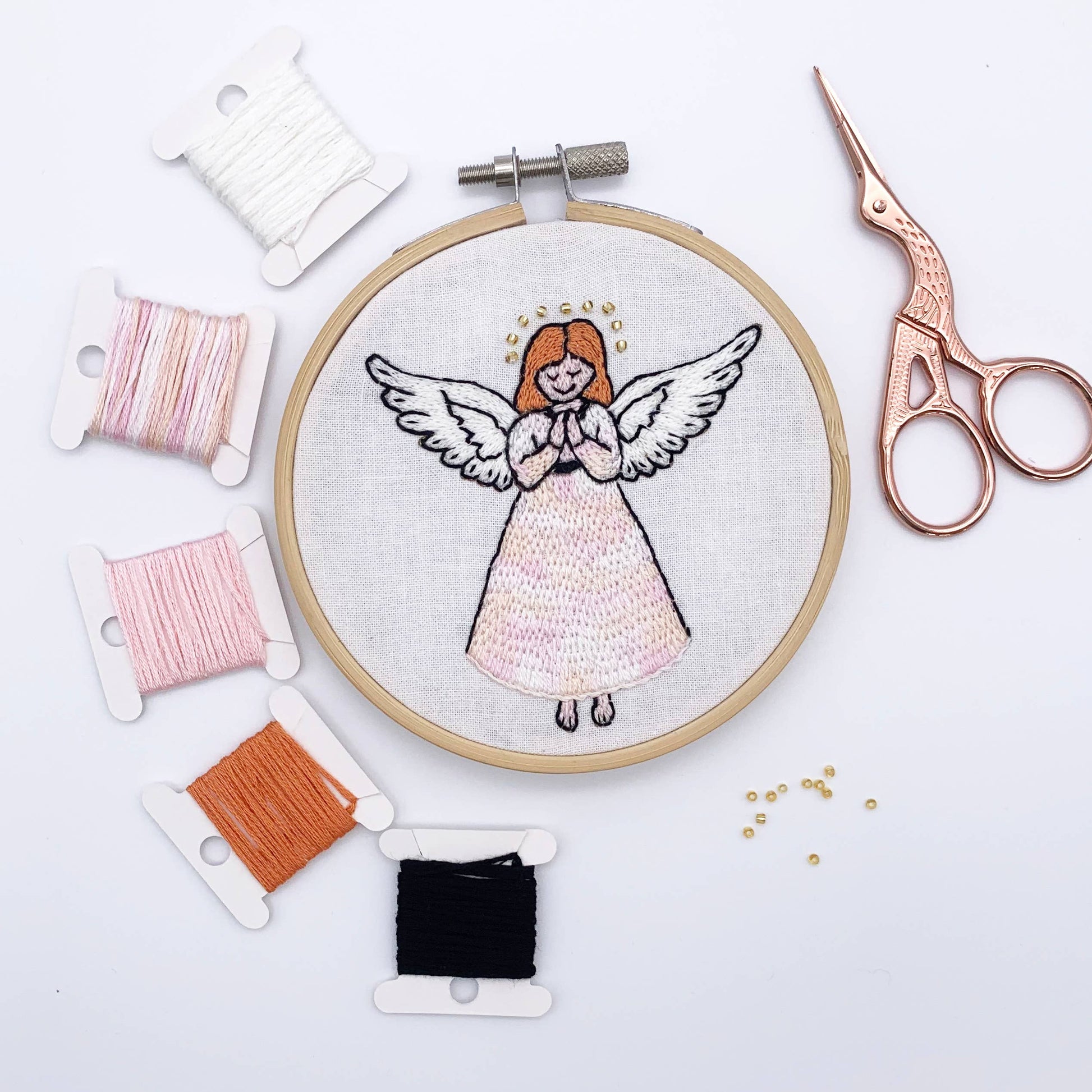 A circular embroidery hoop displaying a finished angel embroidery design on white fabric, surrounded by spools of thread in white, pink, orange, and black, with rose gold embroidery scissors on a white background.