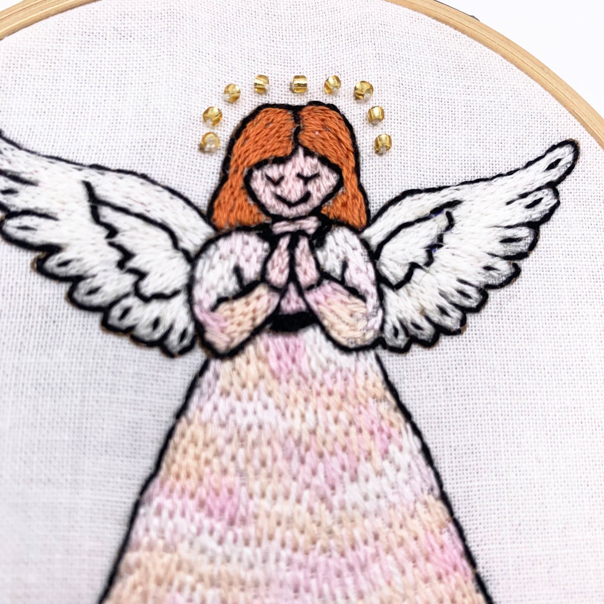 Close-up of a hand-embroidered angel design inside a wooden hoop, featuring white wings, orange hair, and a pink and beige  dress on white fabric.