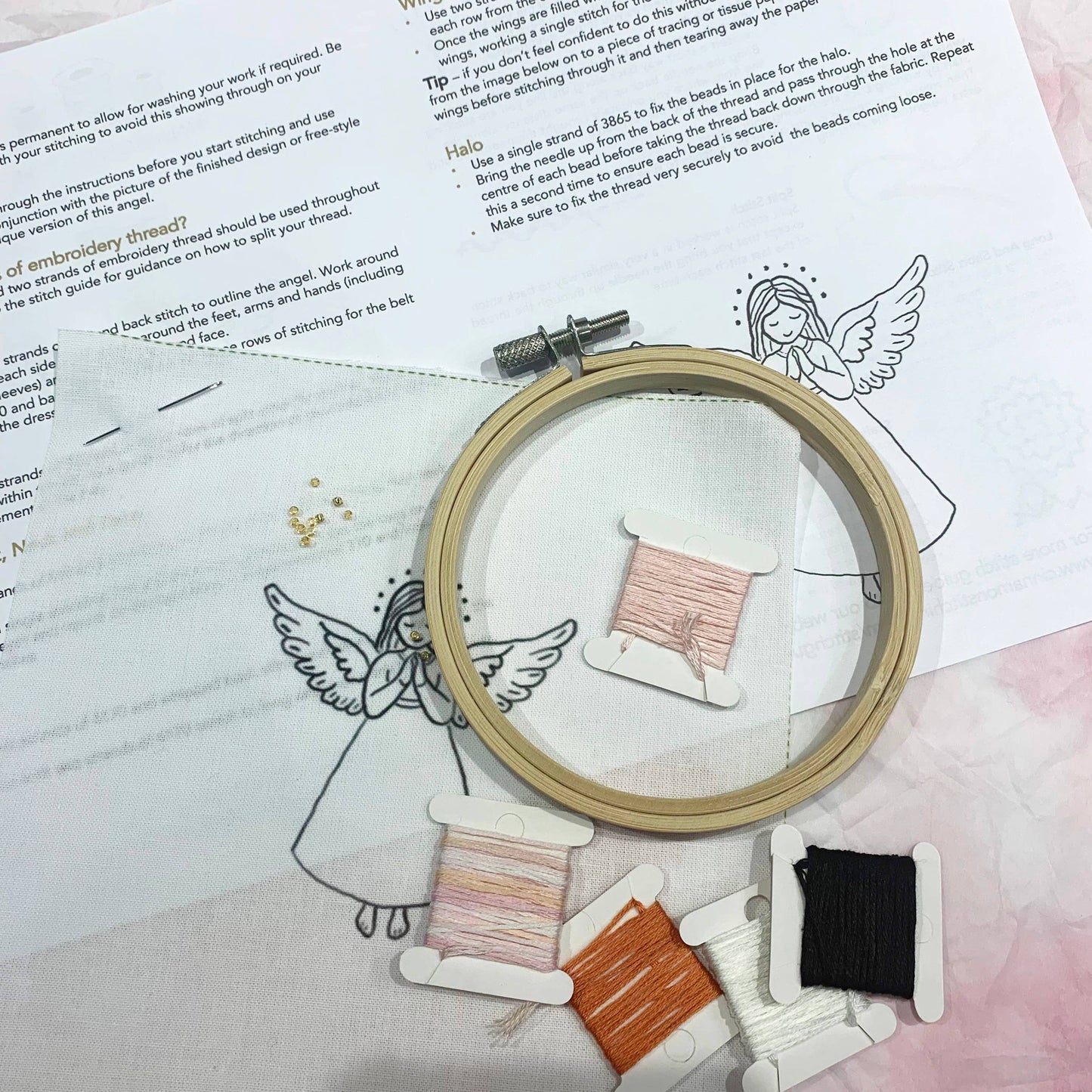 A wooden embroidery hoop containing light pink thread, placed on top of printed embroidery instructions. Several thread spools in white, orange, and black are arranged around the hoop, with small gold beads nearby on a white printed surface.