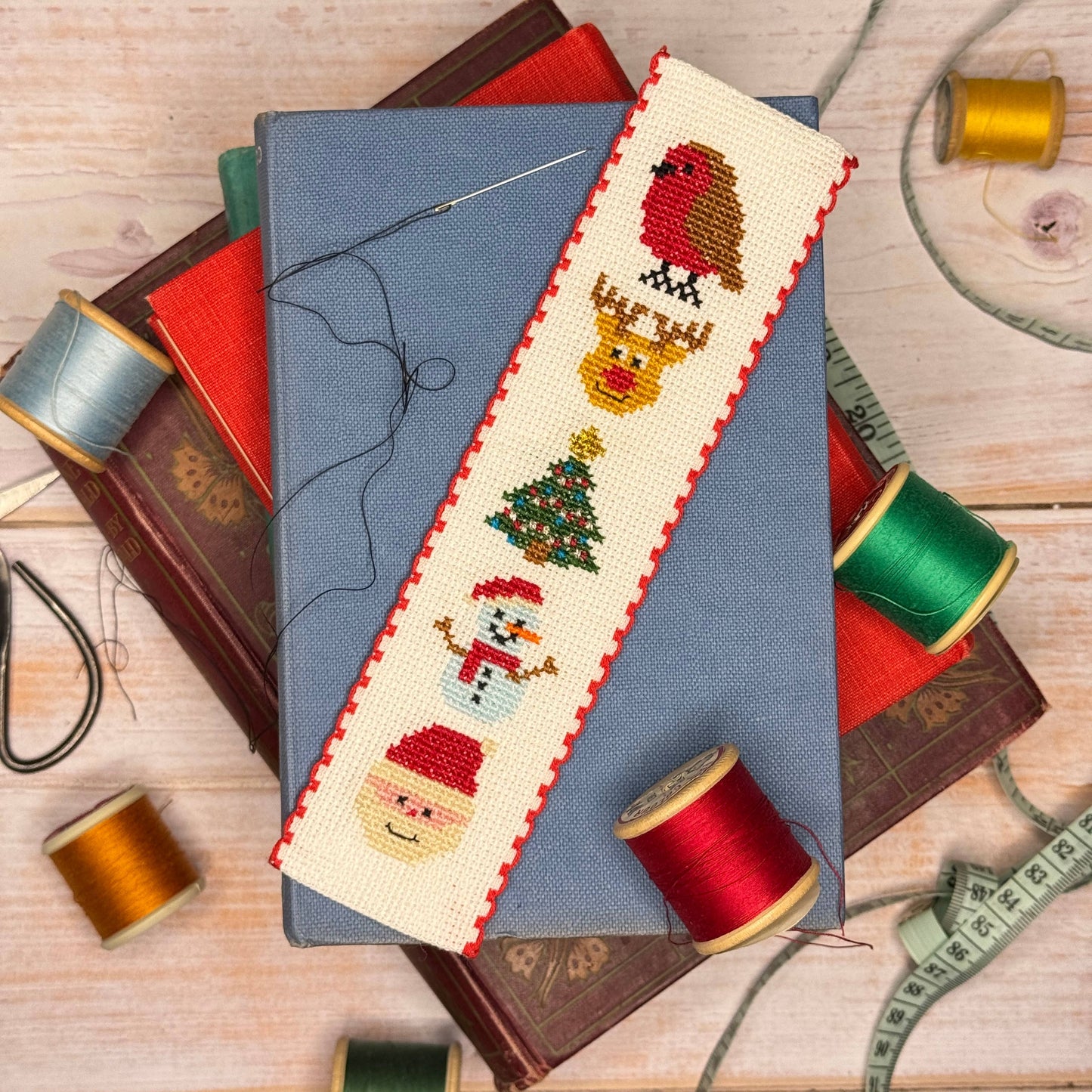 A completed cross-stitch bookmark featuring Christmas characters (robin, reindeer, Christmas tree, snowman, Santa) on  white Aida with red stitched borders, laid diagonally over a blue book surrounded by spools of thread and wooden books on a light wood background.