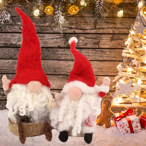 Two festive felt Christmas gnomes with tall red hats and white beards sitting together on a rustic wooden plank surface, with a decorated Christmas tree, warm string lights, pine branches, and wrapped gifts in the background.