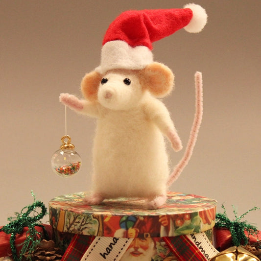 A felt Christmas mouse figurine wearing a red Santa hat, standing on a decorated gift box and holding a small ornament, with holiday decorations around it against a plain beige background.