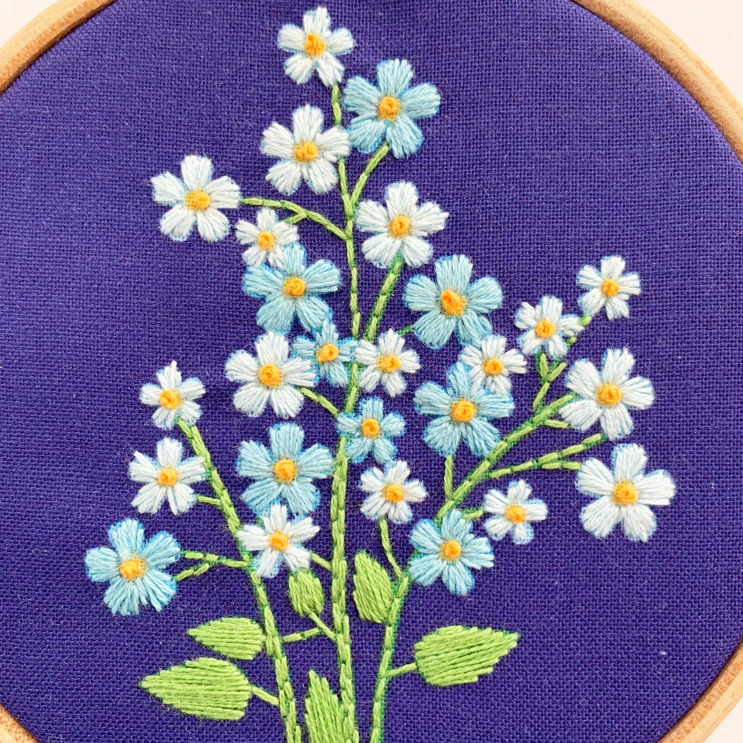 Close-up of a circular embroidery hoop showing a floral design with white and light blue flowers, yellow centers, and green stems on a purple fabric background.
