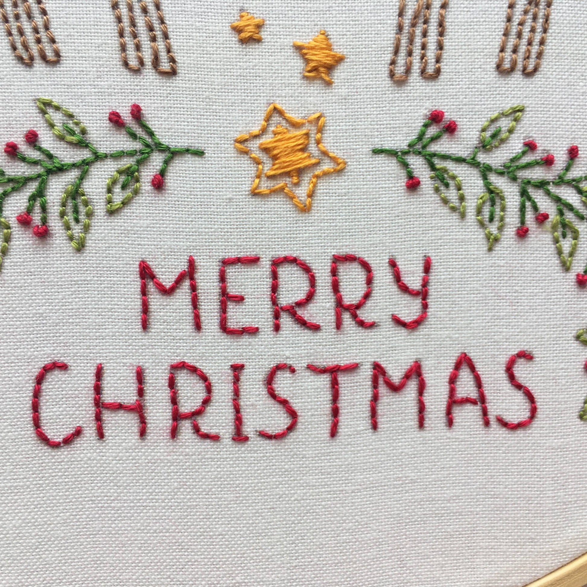 Close-up of a white fabric embroidery hoop featuring the words 'MERRY CHRISTMAS' stitched in red thread, decorated with green holly leaves and red berries, gold stars, and brown decorative elements at the top.