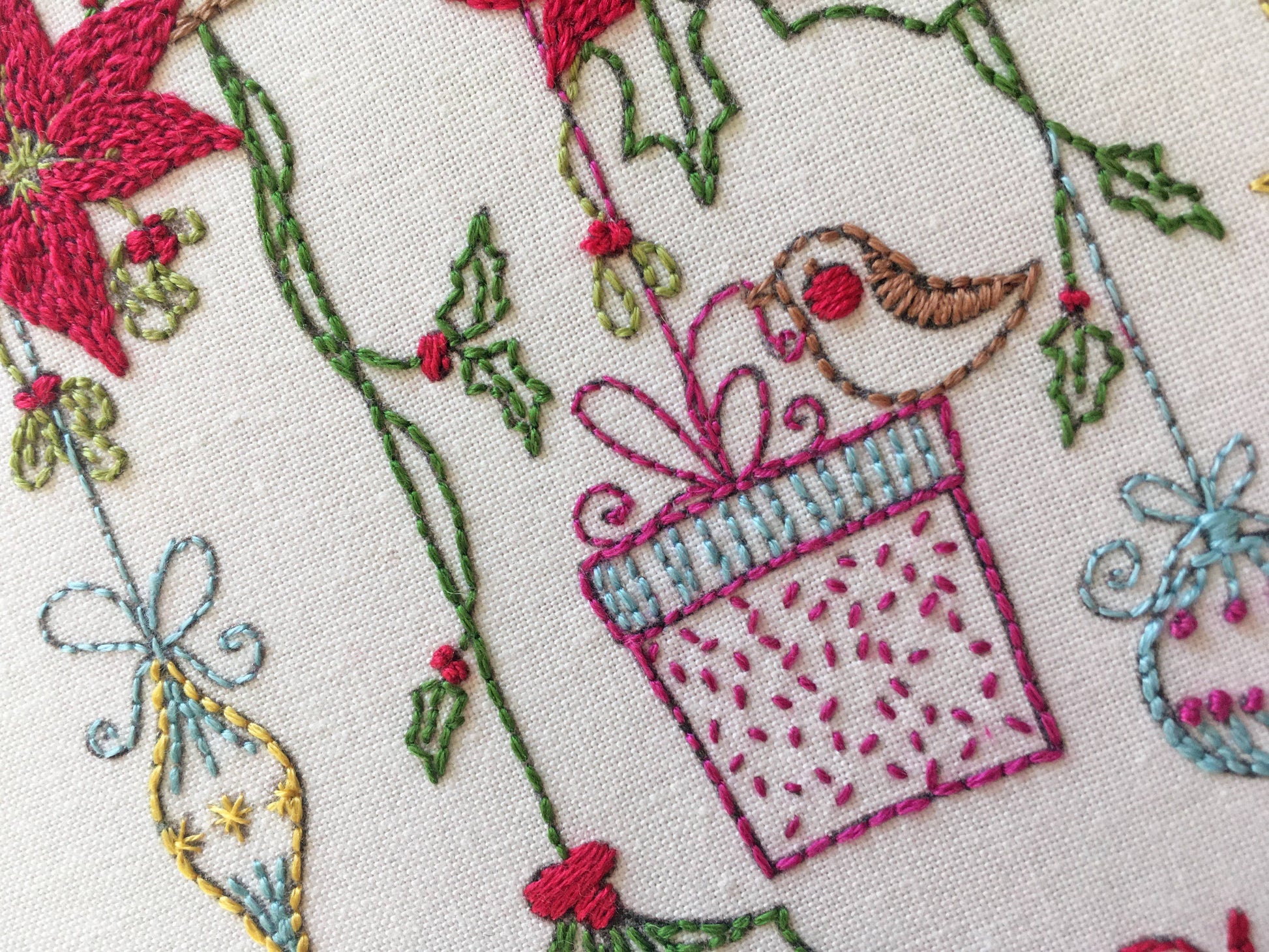 Close-up of a white fabric with colorful embroidered Christmas motifs, including a pink gift box, green holly leaves, red berries, a brown bird, and blue ornaments.