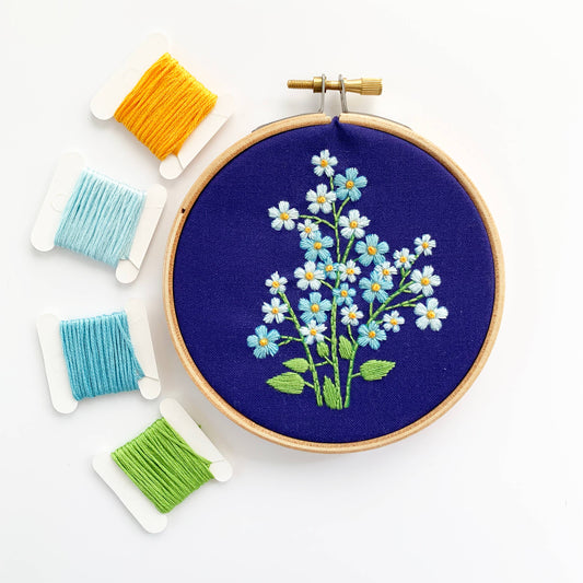 A wooden embroidery hoop on a white background, holding dark blue fabric with a stitched floral design of forget me nots and green stems. Four spools of embroidery thread in yellow, light blue, medium blue, and green are arranged to the left.
