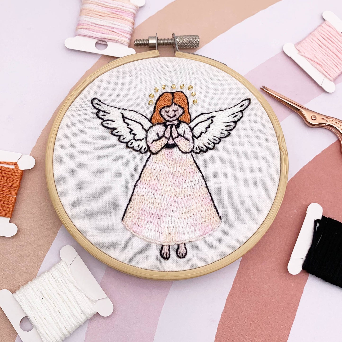 A round embroidery hoop holding white fabric with a stitched angel design, surrounded by spools of thread and embroidery scissors on a pastel pink and beige background.