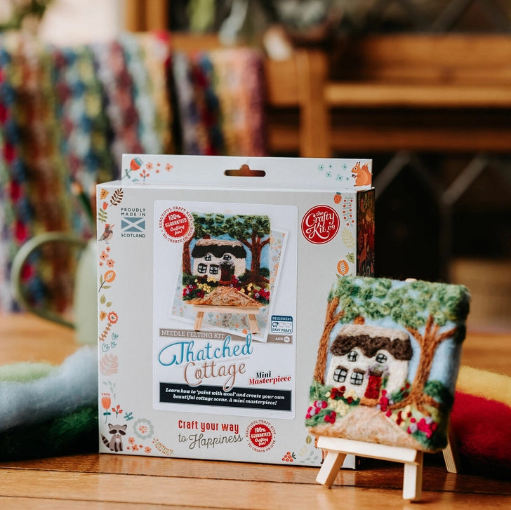A packaged needle felt kit labelled 'Thatched Cottage' stands next to a small finished piece on a wooden tabletop in a real indoor setting with blurred background objects.