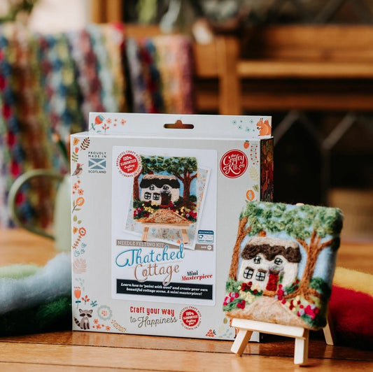 A packaged needle felt kit labelled 'Thatched Cottage' stands next to a small finished piece on a wooden tabletop in a real indoor setting with blurred background objects.