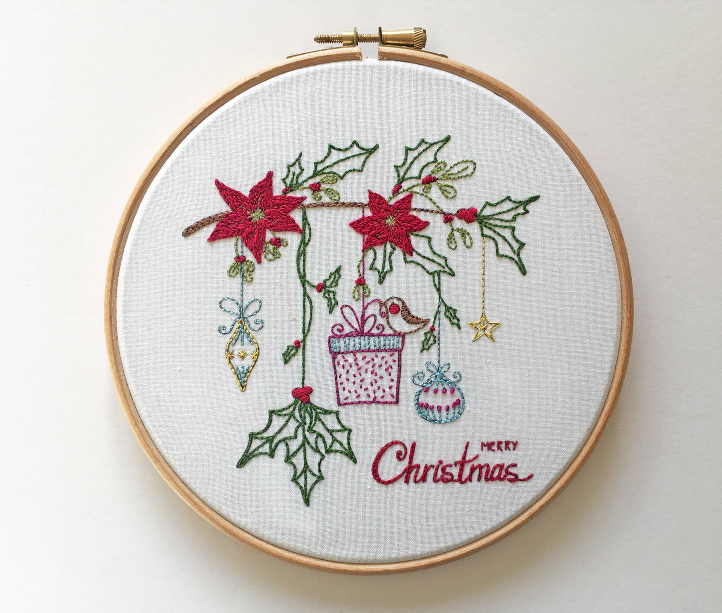 A circular embroidery hoop containing a white fabric backdrop with a Christmas-themed stitched design. The design features red poinsettias, green holly leaves, hanging ornaments, a gift box, and the words 'Merry Christmas' stitched in red script.