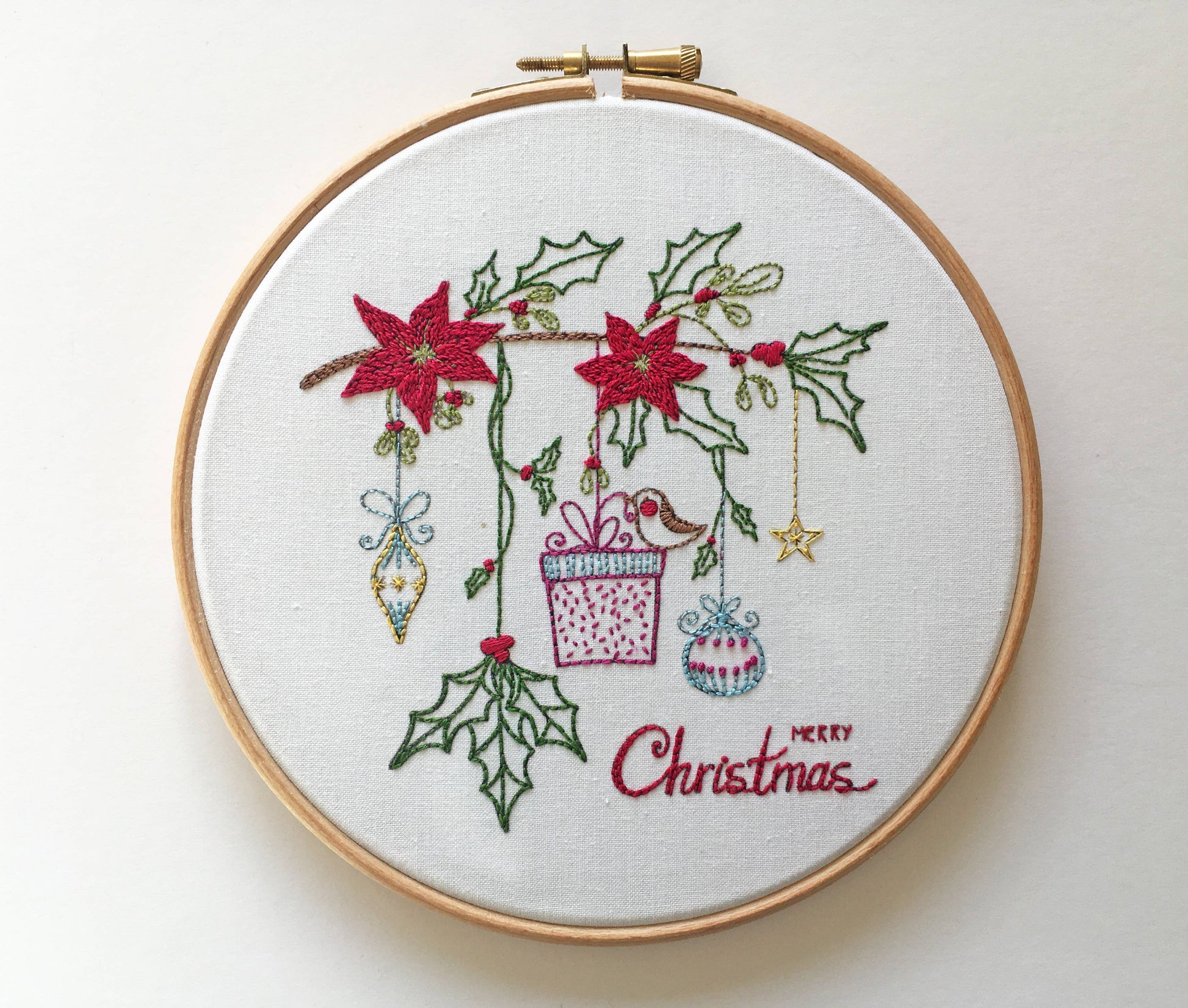 A circular embroidery hoop containing a white fabric backdrop with a Christmas-themed stitched design. The design features red poinsettias, green holly leaves, hanging ornaments, a gift box, and the words 'Merry Christmas' stitched in red script.