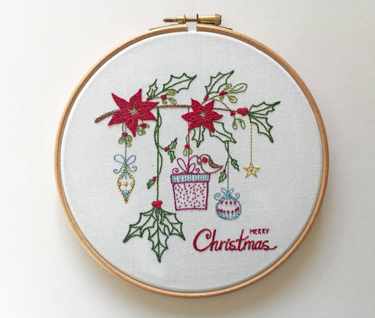 A circular embroidery hoop containing a white fabric backdrop with a Christmas-themed stitched design. The design features red poinsettias, green holly leaves, hanging ornaments, a gift box, and the words 'Merry Christmas' stitched in red script.