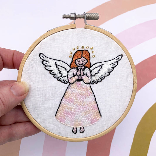 A hand holding a wooden embroidery hoop with a finished embroidered angel design on white fabric. The angel has orange hair, white wings, and a pale pink dress, with a small gold halo of beads.