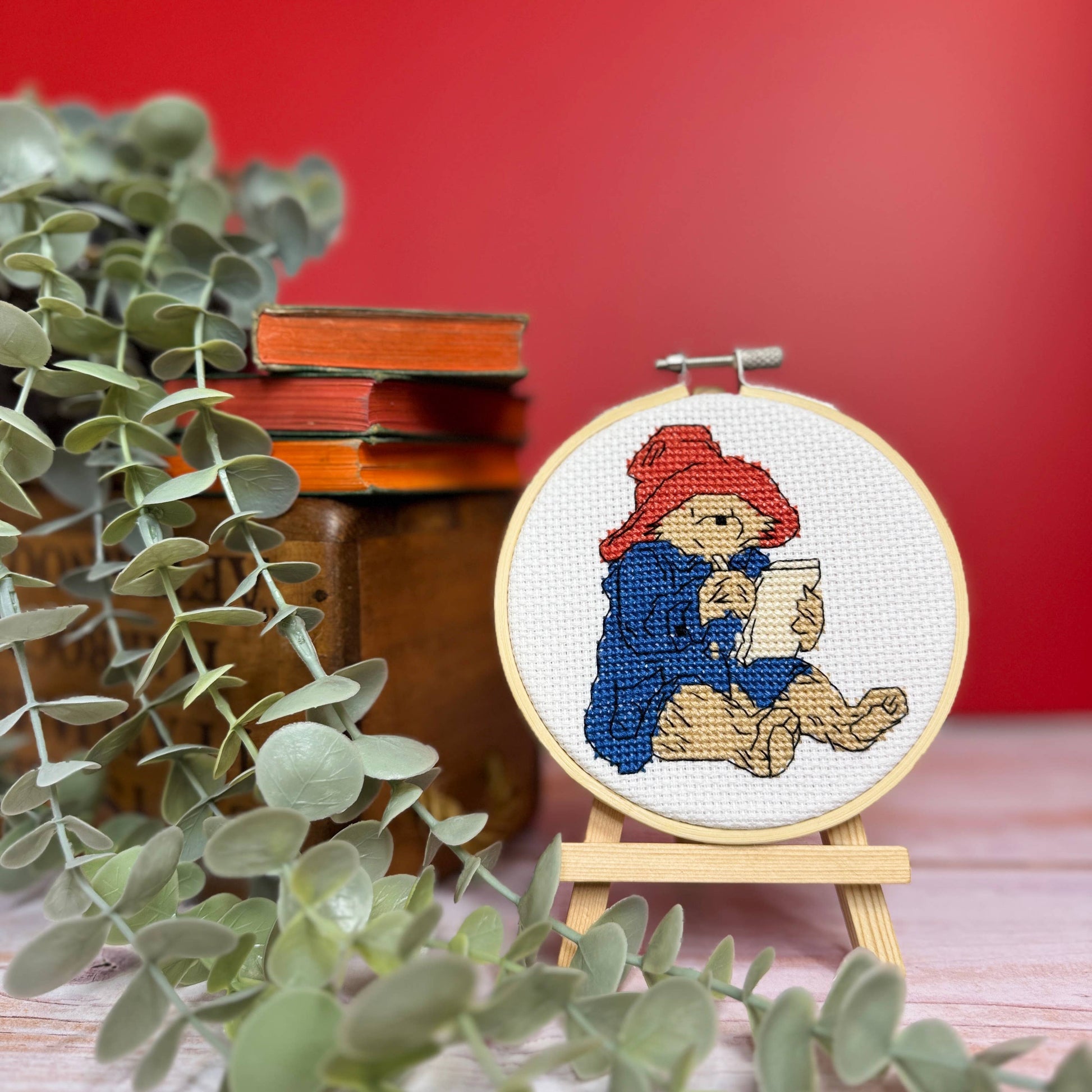 A small round embroidery hoop displaying a cross-stitch design of Paddington Bear writing, set on a miniature stand. The hoop is next to eucalyptus leaves and stacked vintage books against a solid red background.