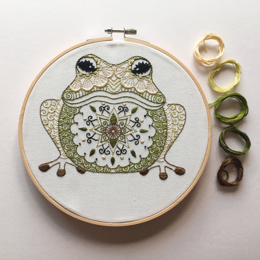 A circular embroidery hoop holding a frog design with ornate green, beige, and white stitching, photographed on a light neutral background. Five rolled threads in yellow, greens, and brown are placed to the right.
