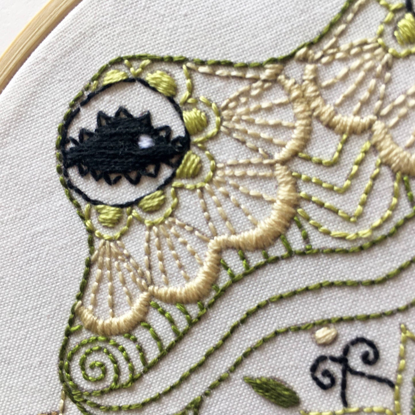 Close-up of an embroidered textile frog design featuring a stylized face with a black eye, cream and olive green stitched patterns, and a light wood hoop edge visible on the left.