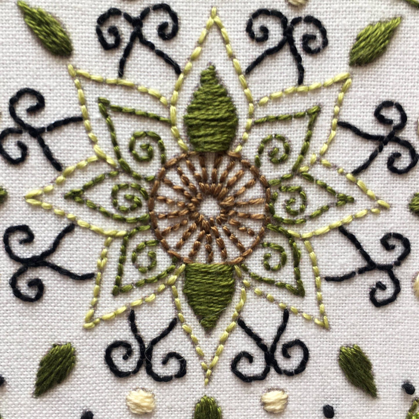 Close-up of a detailed embroidery design on white fabric, featuring a central sunburst motif in brown, olive green, and light yellow, surrounded by black scrollwork and small green leaf accents.