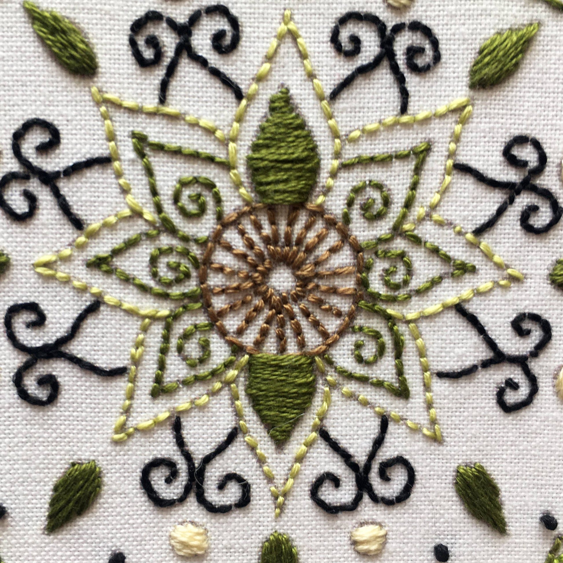 Close-up of a detailed embroidery design on white fabric, featuring a central sunburst motif in brown, olive green, and light yellow, surrounded by black scrollwork and small green leaf accents.