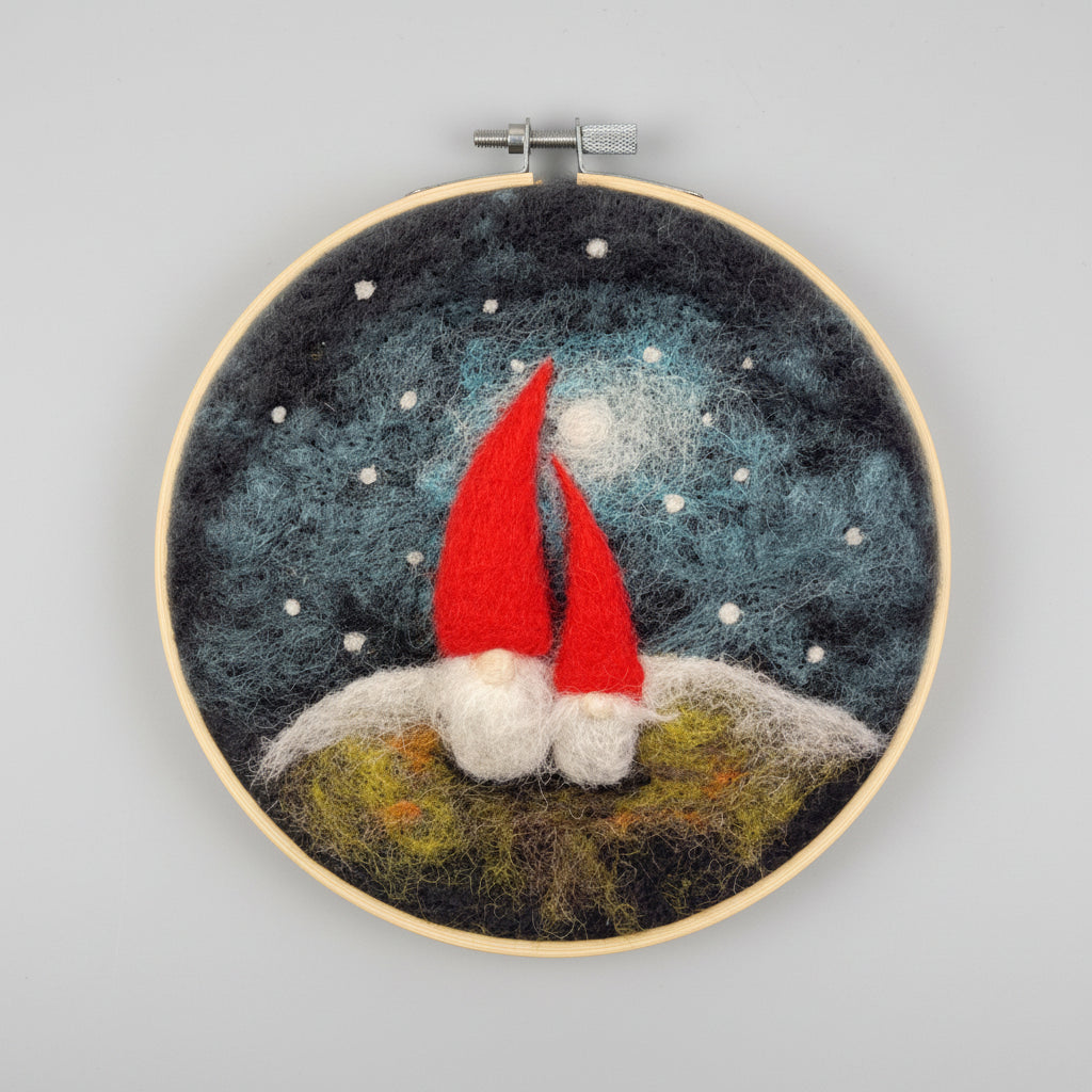 A circular needlepoint embroidery hoop featuring two small gnomes with bright red hats and white beards sitting on a grassy hill under a dark blue starry night sky.