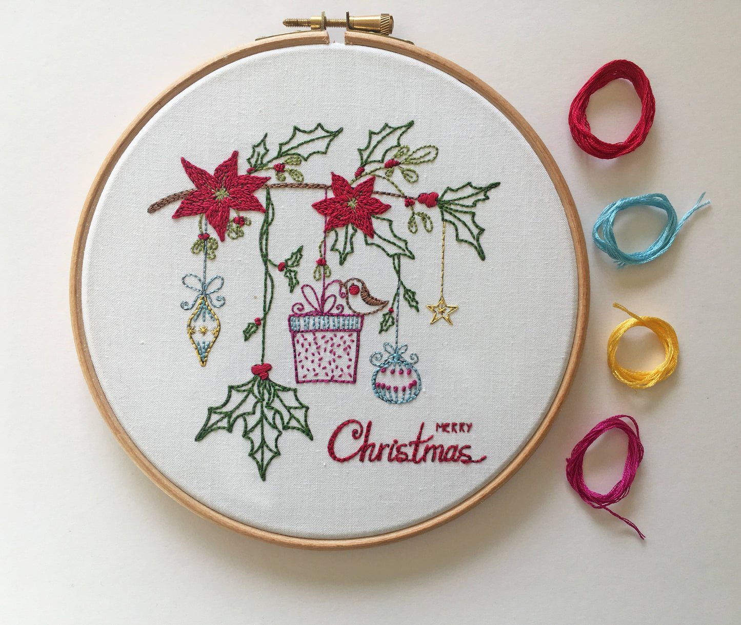 A round embroidery hoop containing a Christmas-themed stitched design with red poinsettias, green holly leaves, ornaments, a gift box, and the words 'Merry Christmas.' Several loose spools of thread in red, blue, yellow, and magenta are arranged to the right on a plain light background.