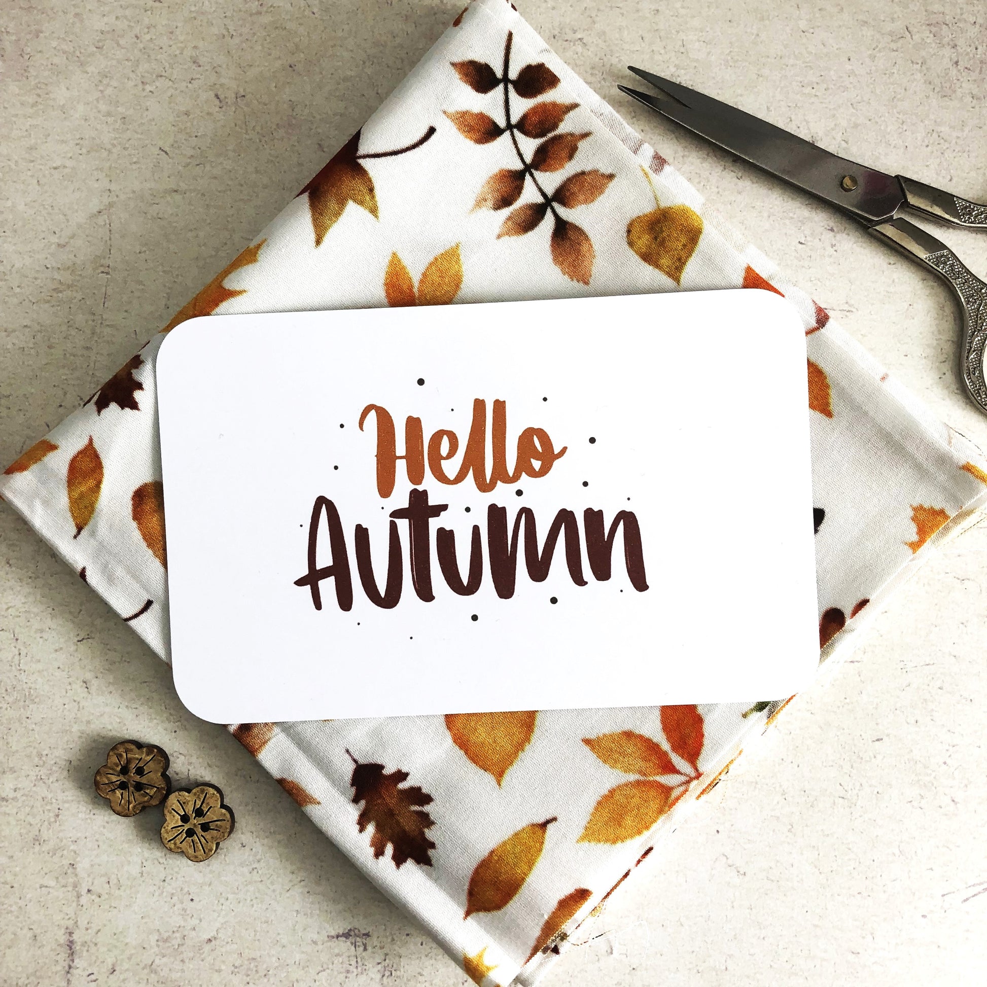 A white rectangular card with the words 'Hello Autumn' in brown lettering, placed on top of folded white fabric printed with autumn leaves in orange, brown, and yellow. Scissors and two wooden buttons are visible as styled props on a light stone surface.