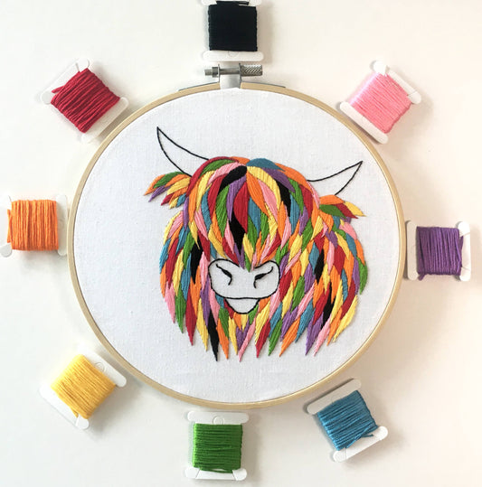 Embroidery hoop with a colourful Highland Cow design surrounded by spools of thread on a white background