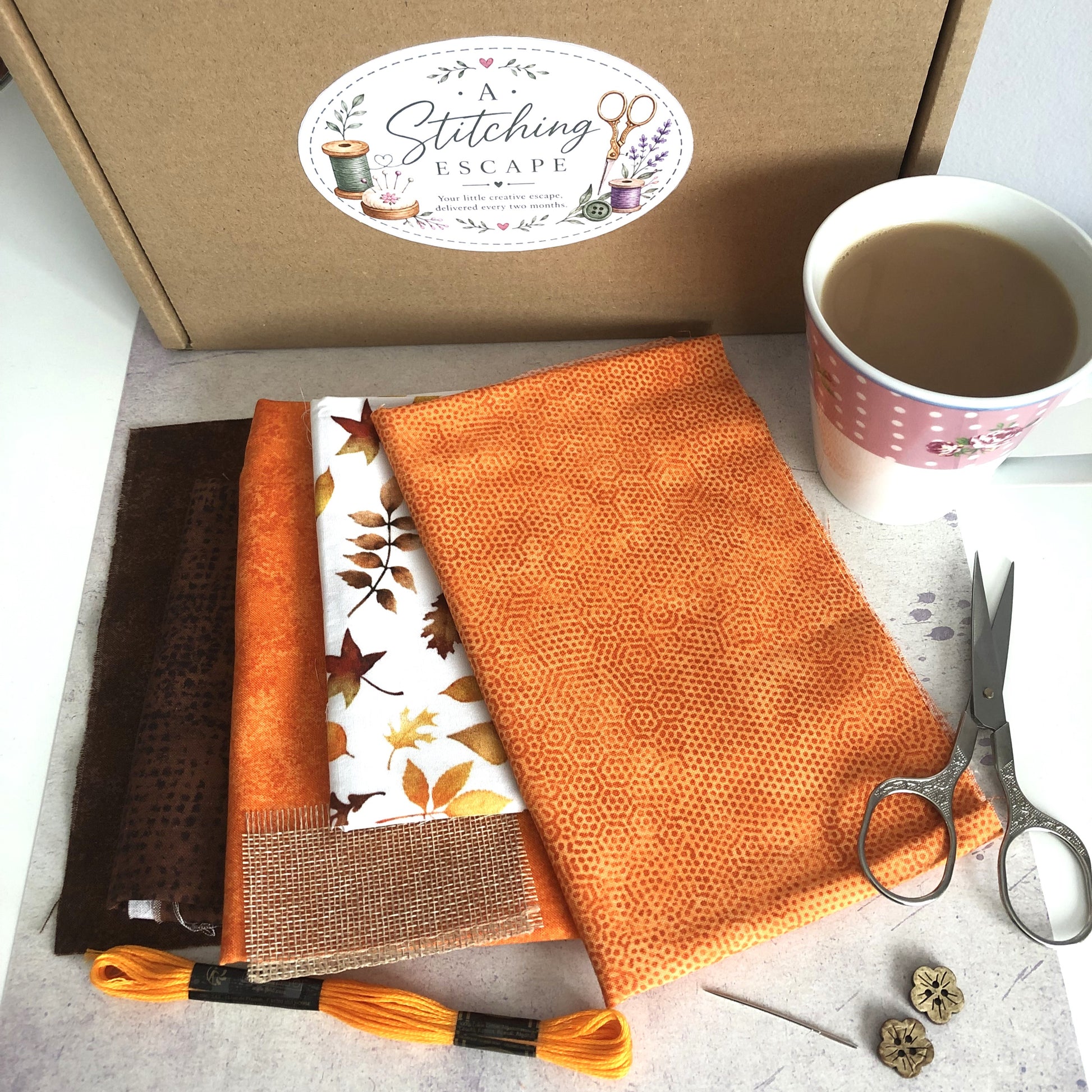 A Stitching Escape craft kit contents arranged on a light surface, including orange and brown fabrics, a white fabric with autumn leaves, embroidery floss, scissors, a needle, buttons, and a branded brown box with a coffee mug nearby.