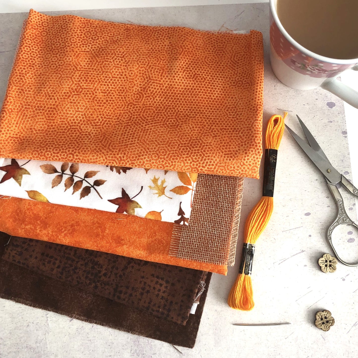 A stack of autumn-themed fabric swatches in orange, brown, and white with leaf patterns, displayed with embroidery floss, scissors, a needle, and a coffee mug on a light stone surface.