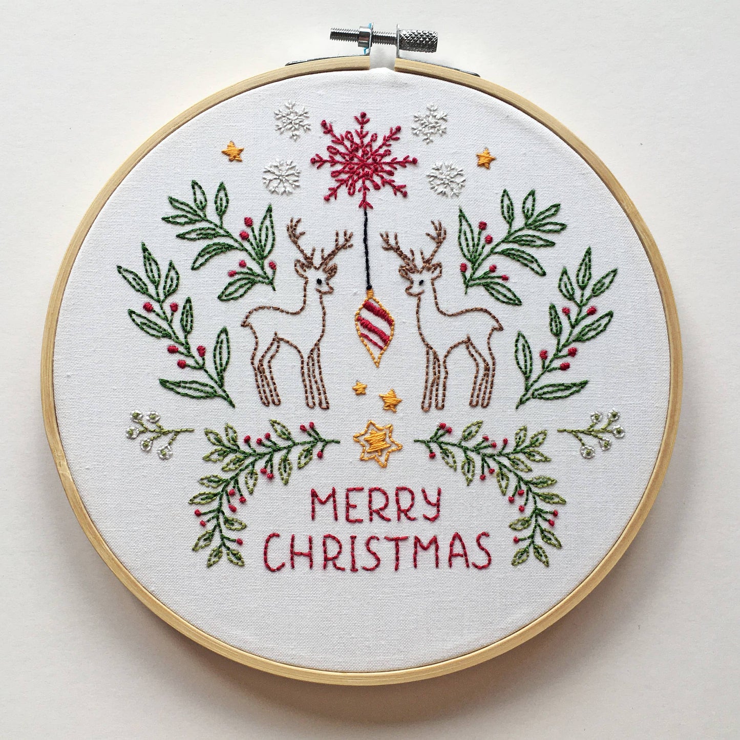 A round embroidery hoop holding a white fabric with a Christmas-themed stitched design. The design features two brown deer, a red ornament, green leafy branches with red berries, small gold stars, white snowflakes, and the words 'MERRY CHRISTMAS' in red.