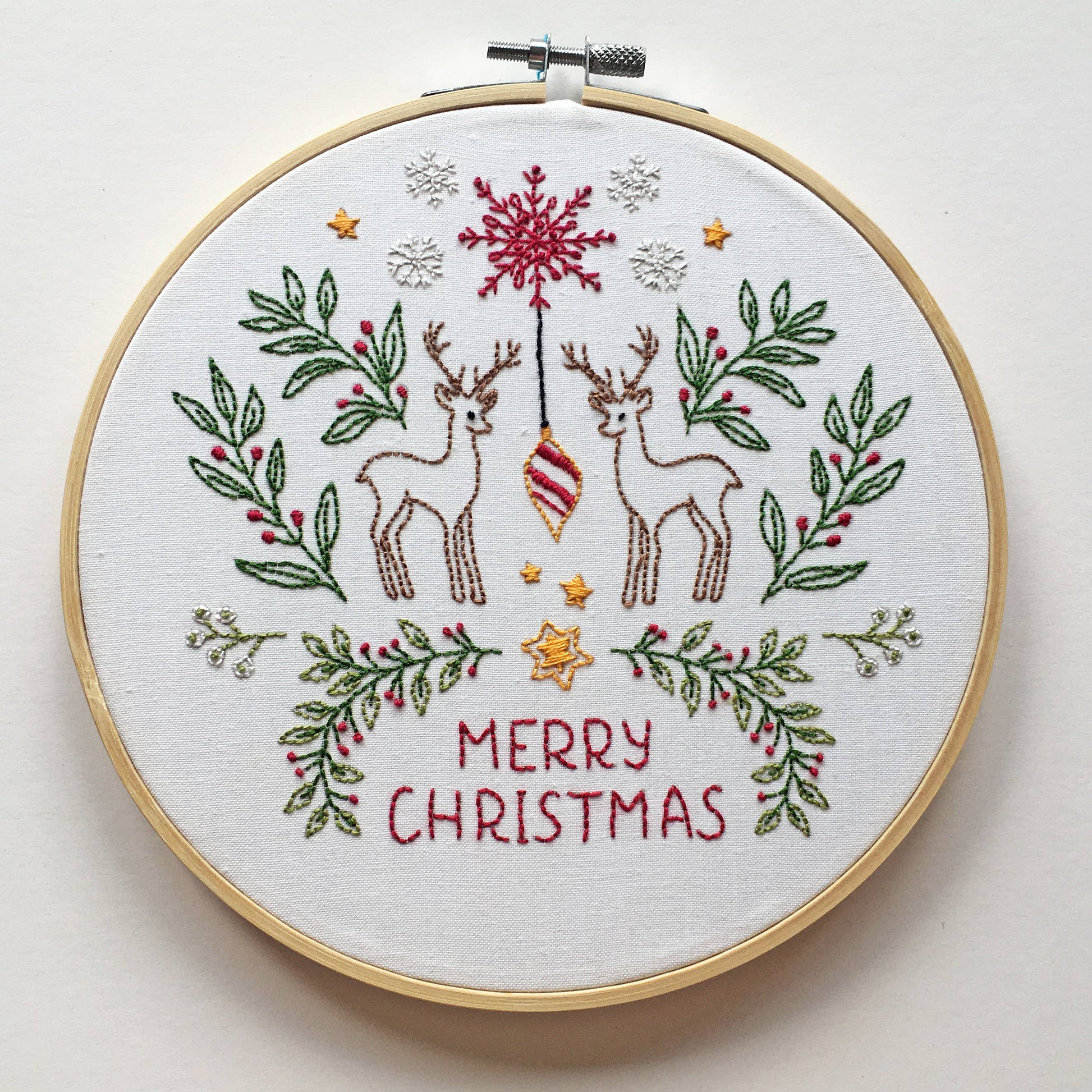 A round embroidery hoop holding a white fabric with a Christmas-themed stitched design. The design features two brown deer, a red ornament, green leafy branches with red berries, small gold stars, white snowflakes, and the words 'MERRY CHRISTMAS' in red.