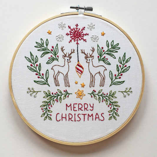 A round embroidery hoop holding a white fabric with a Christmas-themed stitched design. The design features two brown deer, a red ornament, green leafy branches with red berries, small gold stars, white snowflakes, and the words 'MERRY CHRISTMAS' in red.
