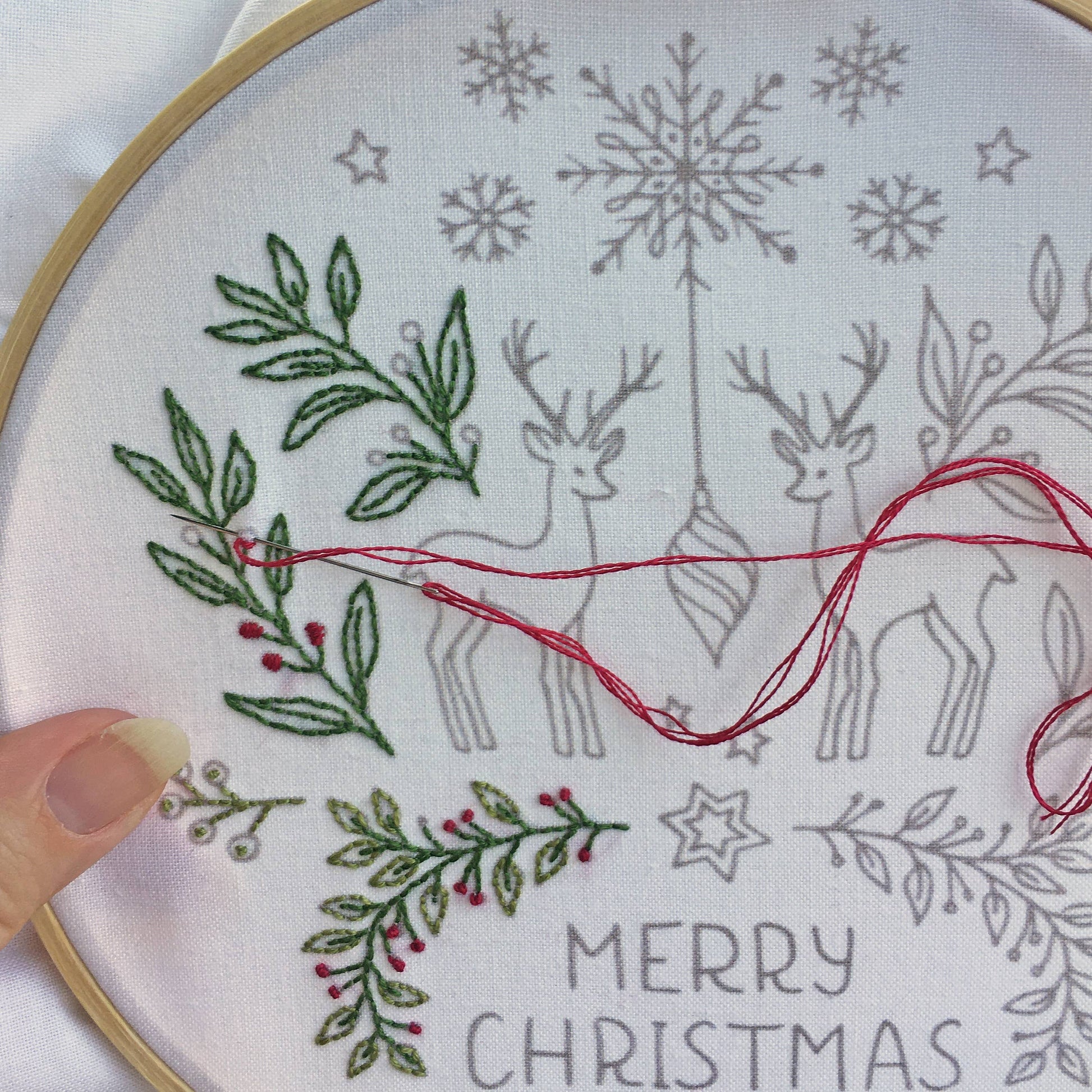 A close-up of a circular embroidery hoop showing a white fabric with gray outlined holiday motifs, including snowflakes, deer, stars, and the words “MERRY CHRISTMAS.” Green leafy branches and small red berry accents are stitched on the left side, with red thread draped across the center.