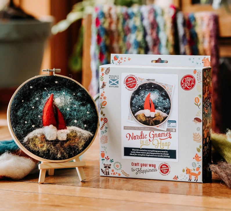 A finished circular embroidery piece showing two gnomes under a snowy night sky, displayed next to its retail packaging box on a wooden table.