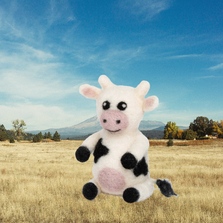 Needle felted cow in a field with a mountain and blue sky in the background