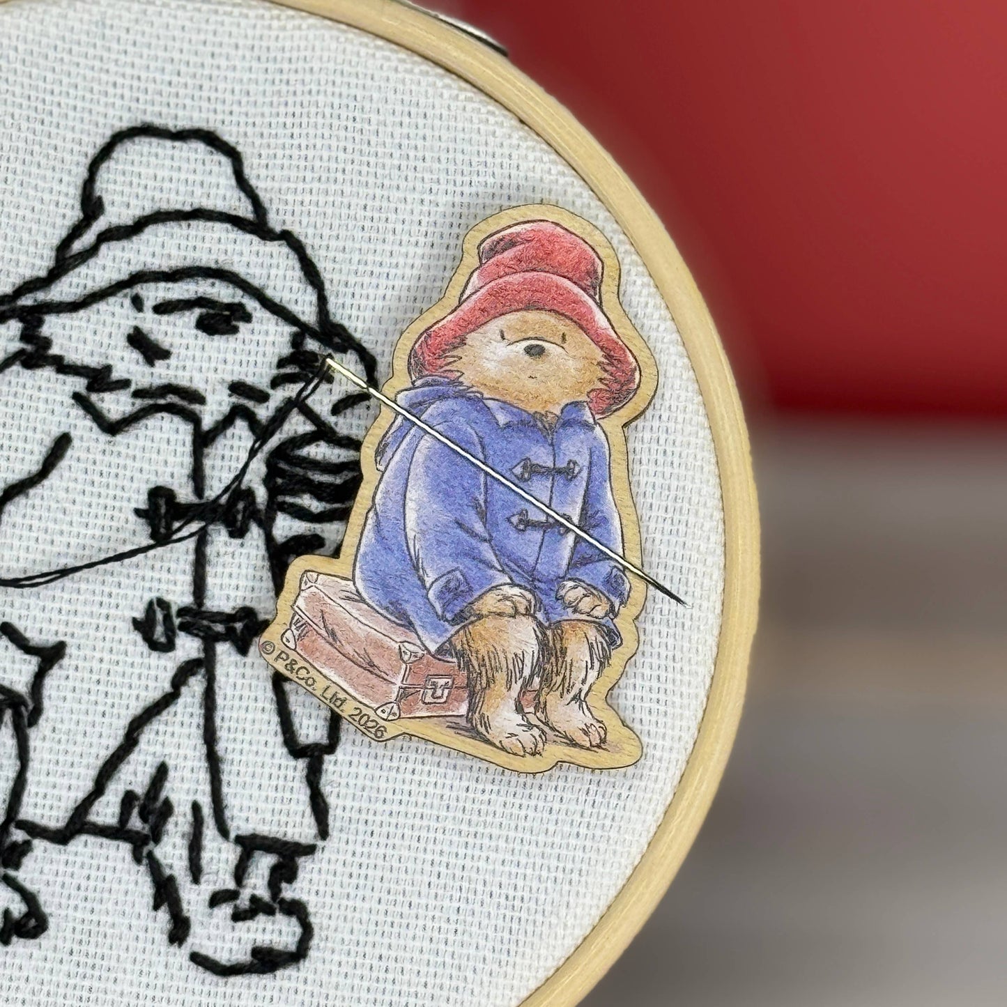 A close-up of a circular embroidery hoop showing a partially stitched black-and-white figure on white fabric, with a needle minder of Paddington Bear pinned over the embroidery.