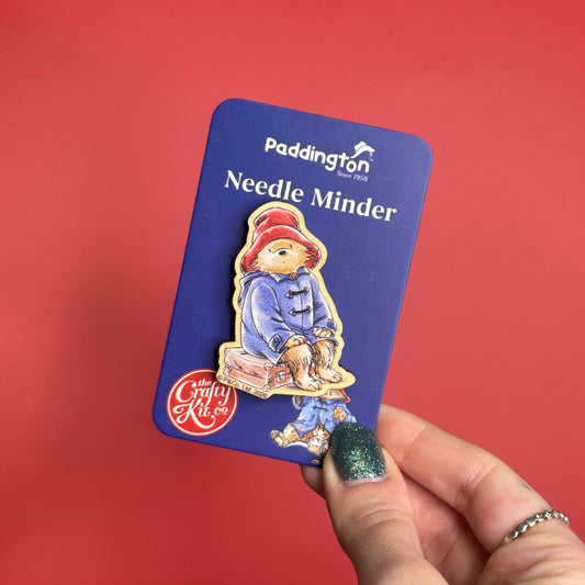 A hand with glittery green nail polish holds a blue card featuring a Paddington-themed enamel pin labeled 'Paddington Needle Minder' and 'The Great Rabbit Co.' against a solid red background.