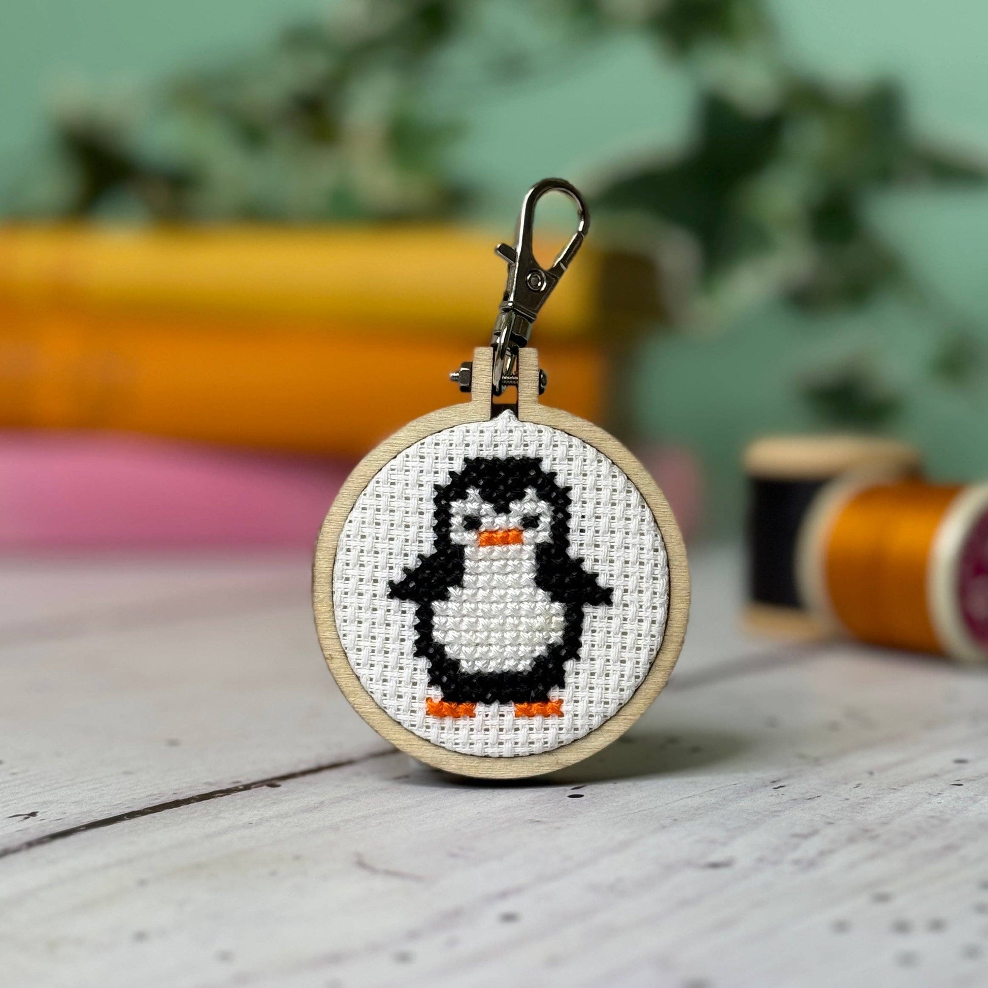 Hoop with a cross stitched design of a penguin with a blurred background of books and sewing reels.