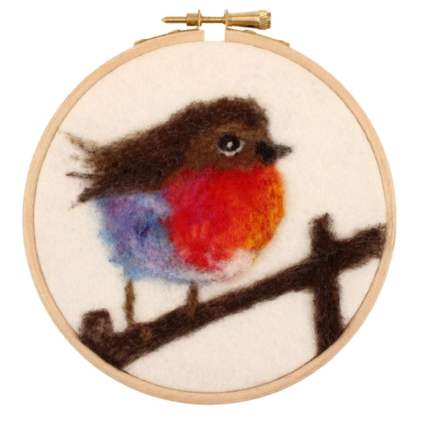 A circular needlepoint embroidery hoop featuring a felted bird with a brown body, bright red-orange chest, and blue-purple wings, perched on a dark brown branch against a light cream background.
