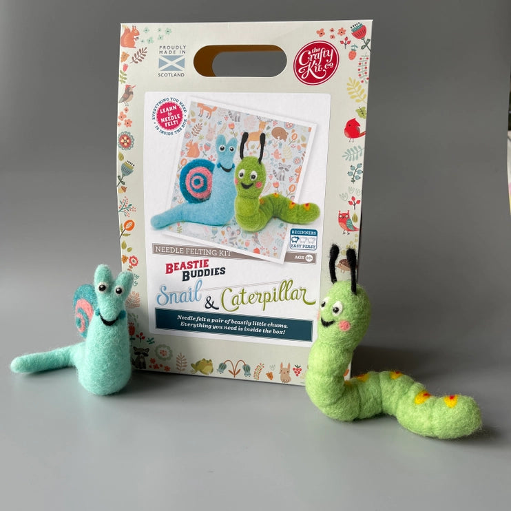 Two needle felted toys, a snail and a caterpillar, in front of their craft kit packaging on a grey background.