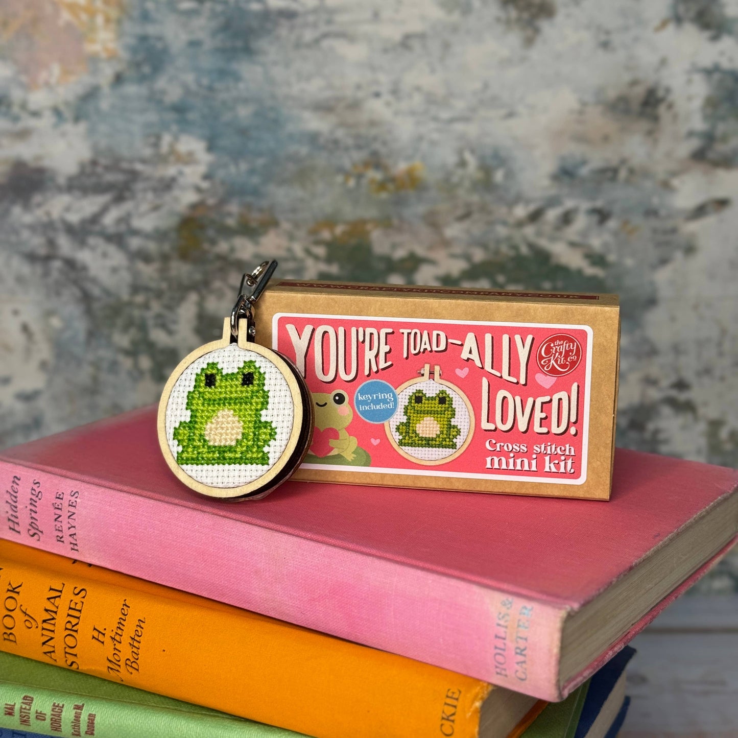 Cross stitch mini kit with a toad design on a stack of books against a textured background