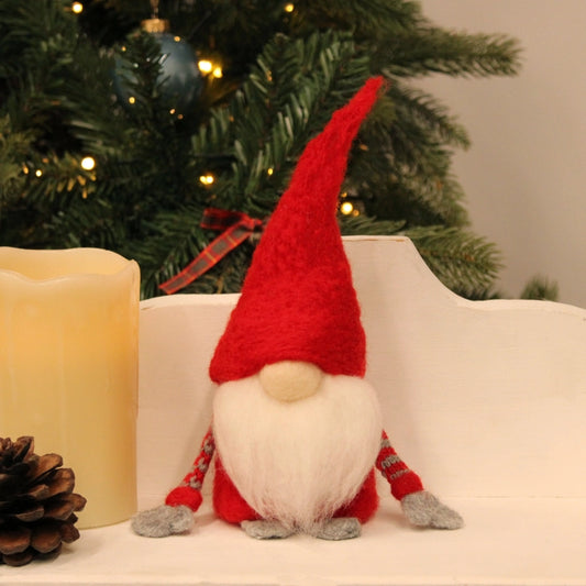 A small Christmas gnome  with a tall red hat, white beard, and red-and-white striped legs sits on a white shelf in front of a decorated Christmas tree. A cream candle and a pinecone are partly visible beside it.